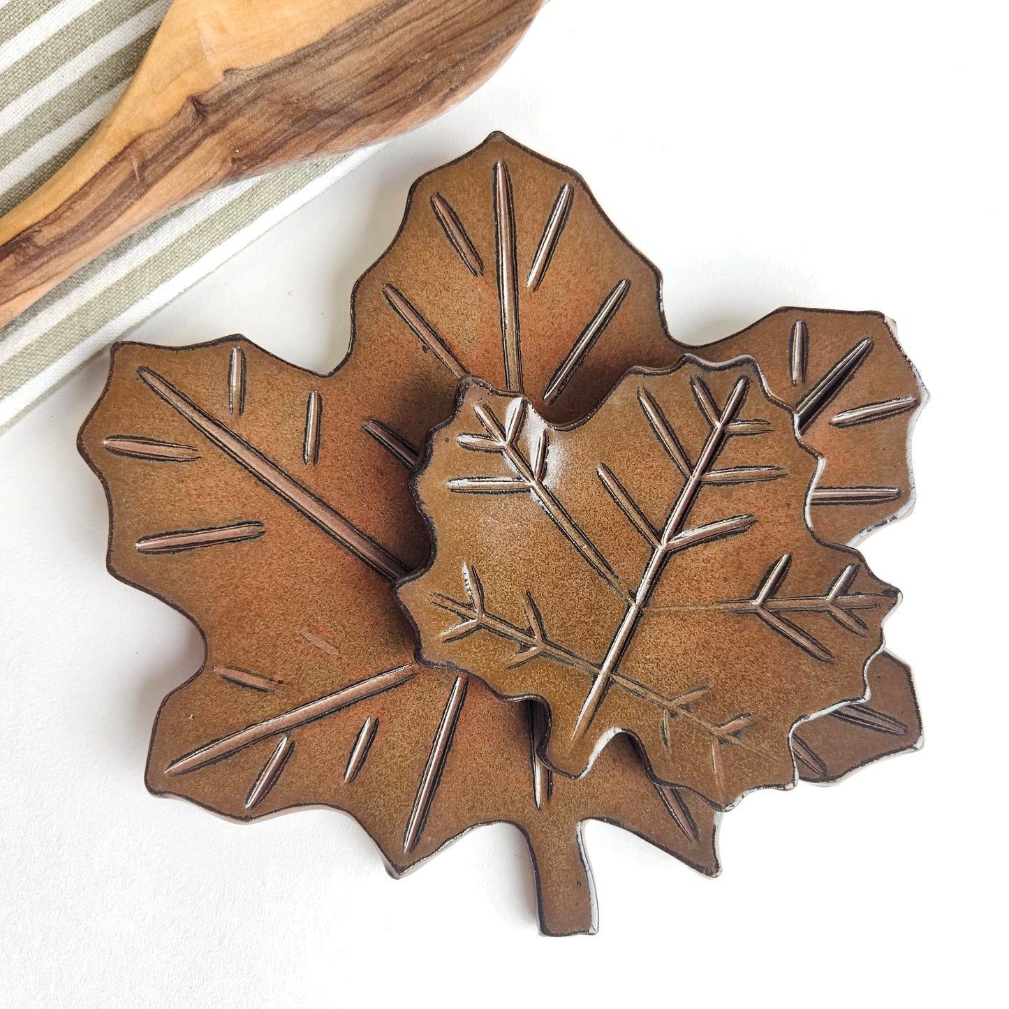 Handmade ceramic leaf-shaped countertop spoon rest on a white background with wooden spoon and cotton kitchen towel nearby