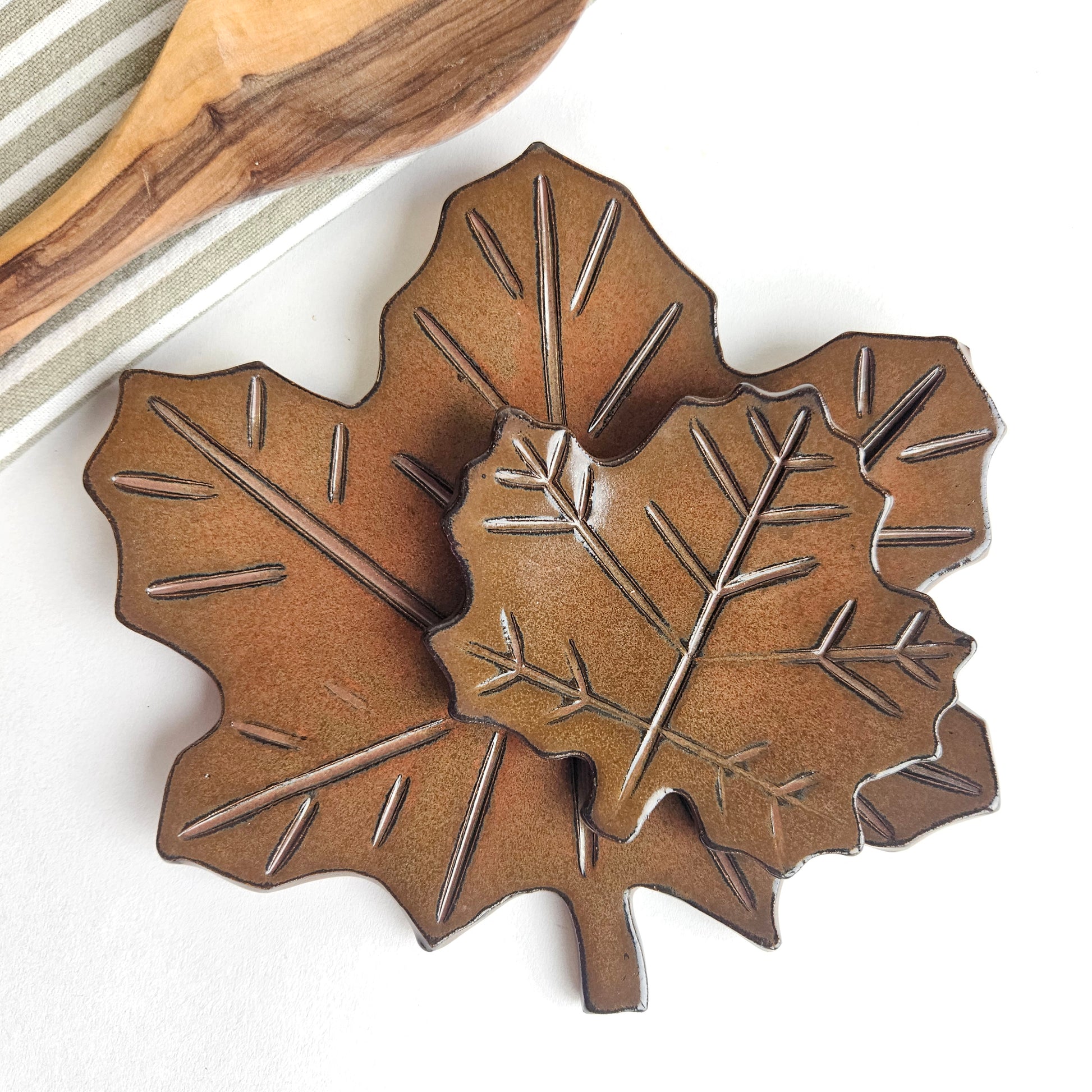 Handmade ceramic leaf-shaped countertop spoon rest on a white background with wooden spoon and cotton kitchen towel nearby