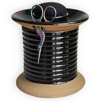 Black ceramic sewing station caddy shaped like an oversized spool with plain black pincushion topper, rainbow crane thread snips with built-in compartments for embroidery sewing and quilting supplies.