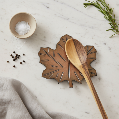Ceramic leaf-shaped trivet with a wooden spoon on a marble surface with cooking herbs and dish towel nearby.