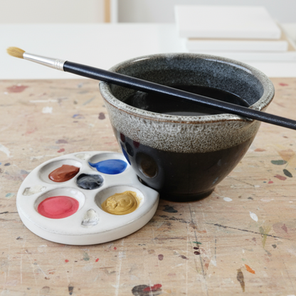 Black and white handmade ceramic watercolor palette and rinse bowl set with paintbrush, displayed on an artist’s worktable.