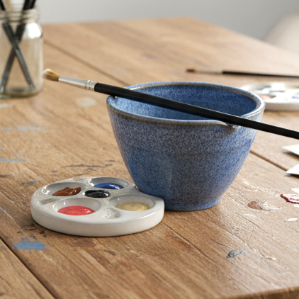 Blue ceramic bowl with paintbrushes on a wooden table next to a paint palette with colors.