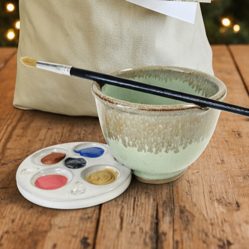 Ceramic paint Rinse bowl with paintbrush and watercolor palette on a wooden surface with canvas gift bag in background