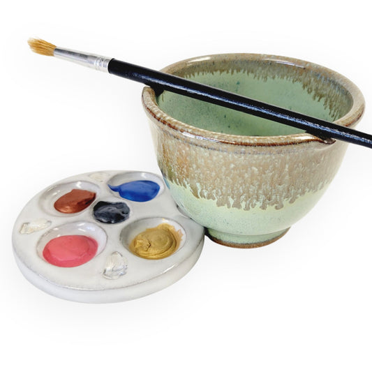 Compact green and bronze ceramic paint palette and brush rinse bowl set with textured wells and built-in brush rest holding blobs of paint and a brush in green and brown glaze.