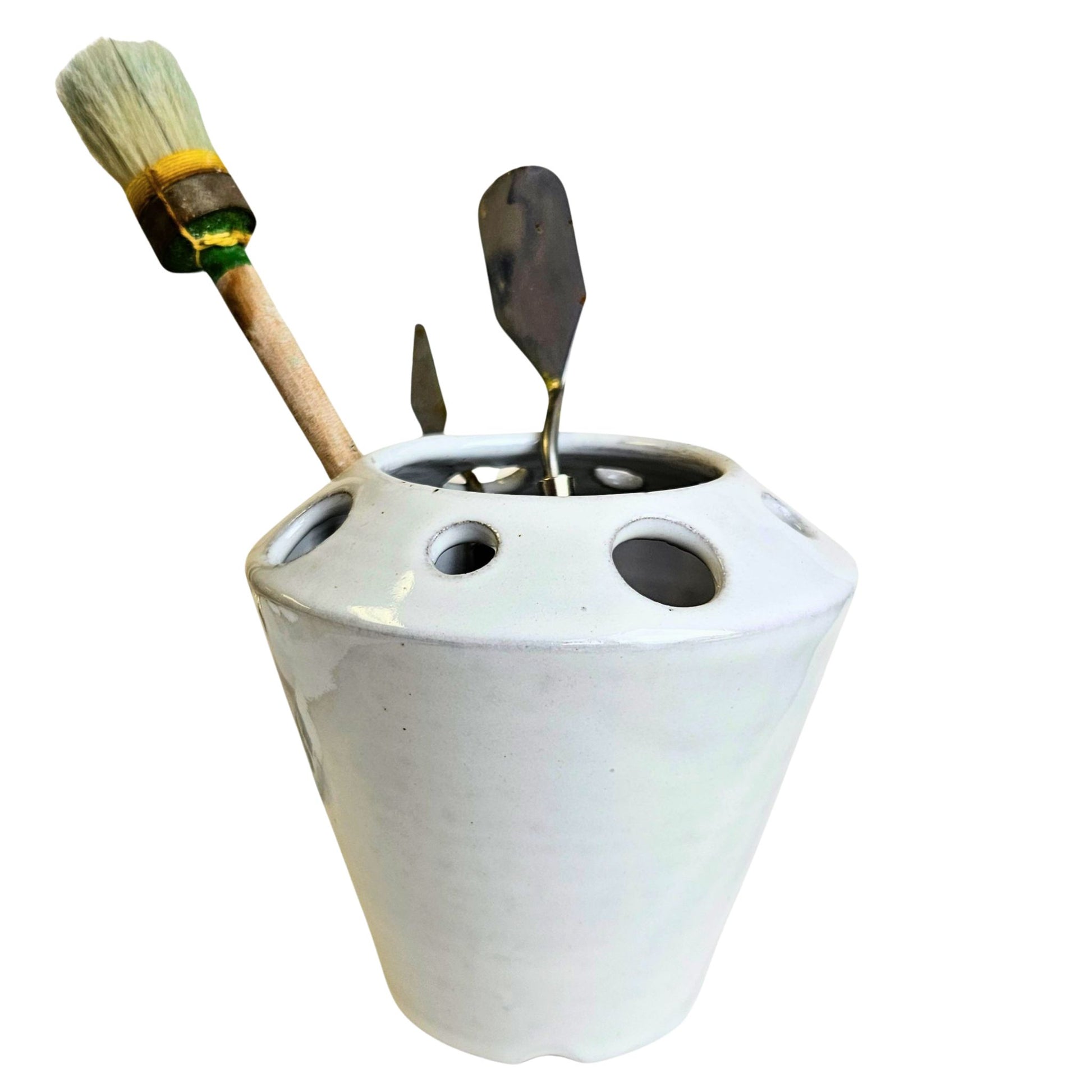 White ceramic craft tool holder with various sized holes containing a paint brush and two palette knives on a white background