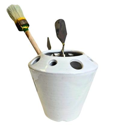 White ceramic craft tool holder with various sized holes containing a paint brush and two palette knives on a white background