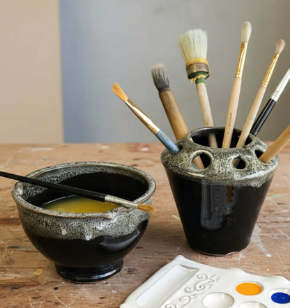 A paintbrush holder and matching paint  rinse cup on a wooden table, with several paintbrushes stored in the holder and a white paint palette visible in the foreground.