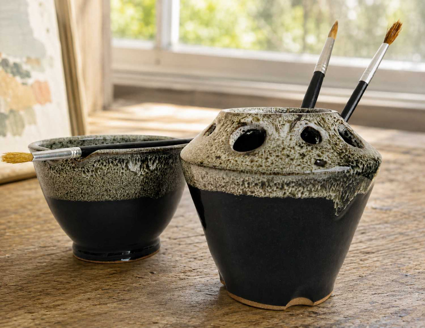 Two paintbrush holders on a wooden table near a window. The larger holder on the right has a dark black and brown speckled glaze with a light beige rim, and two paintbrushes are tucked into its holes.