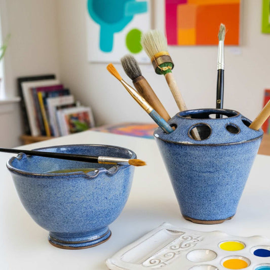A pair of blue ceramic paint storage containers on a white table, one acting as a water bowl with a brush resting across the rim, and the other holding several paintbrushes. A white paint palette is visible in the foreground, with a colorful art room background.