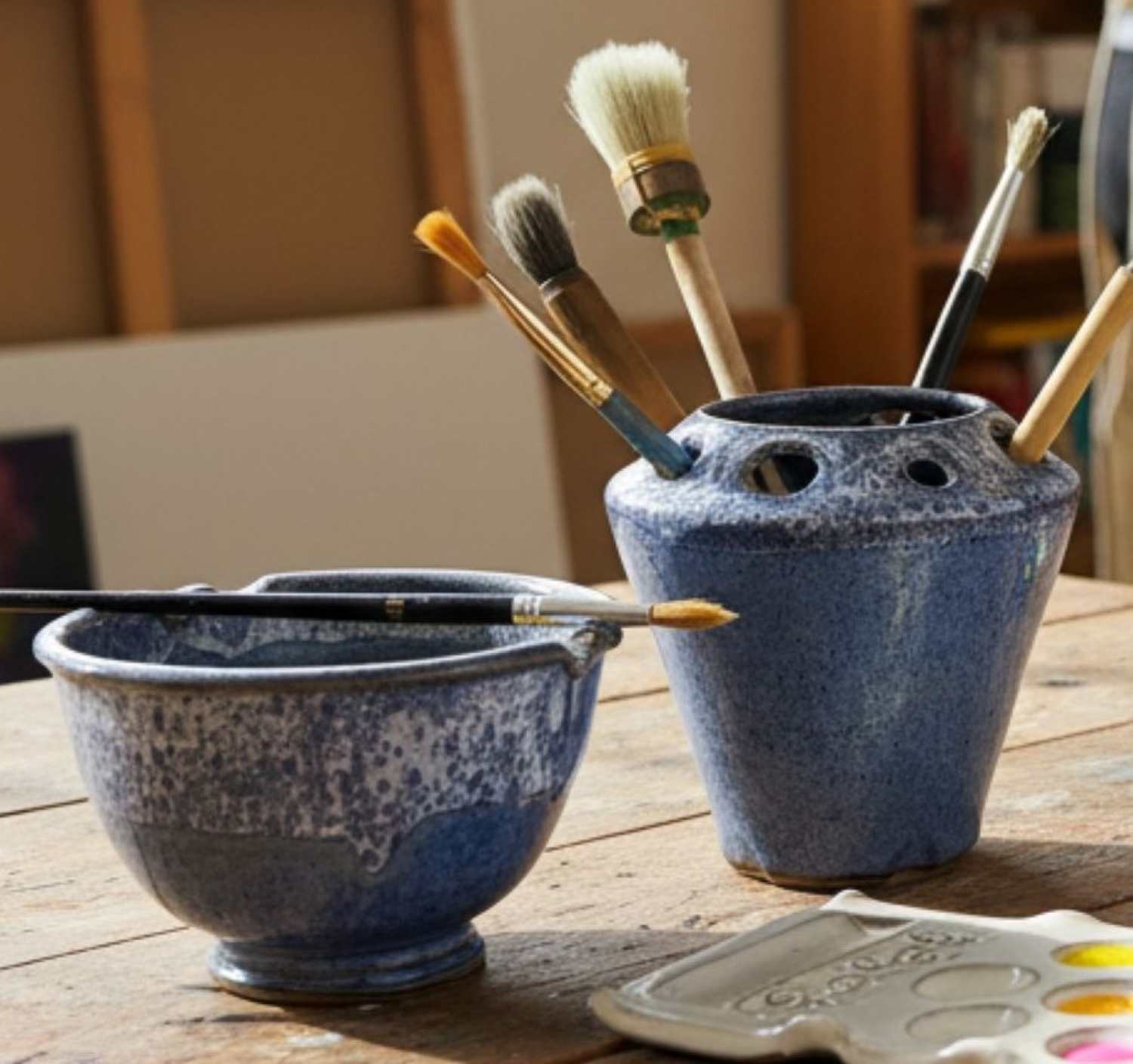A set of painting tools including two blue- ceramic matching pieces -a paint rinse cup, a brush holder, and paintbrushes, arranged on a rustic wooden table next to a paint palette.
