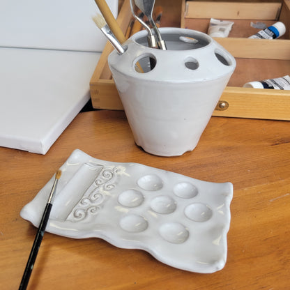 A handcrafted ceramic artist watercolor set including a paint palette, a brush caddy = in white color, displayed on a wooden surface.