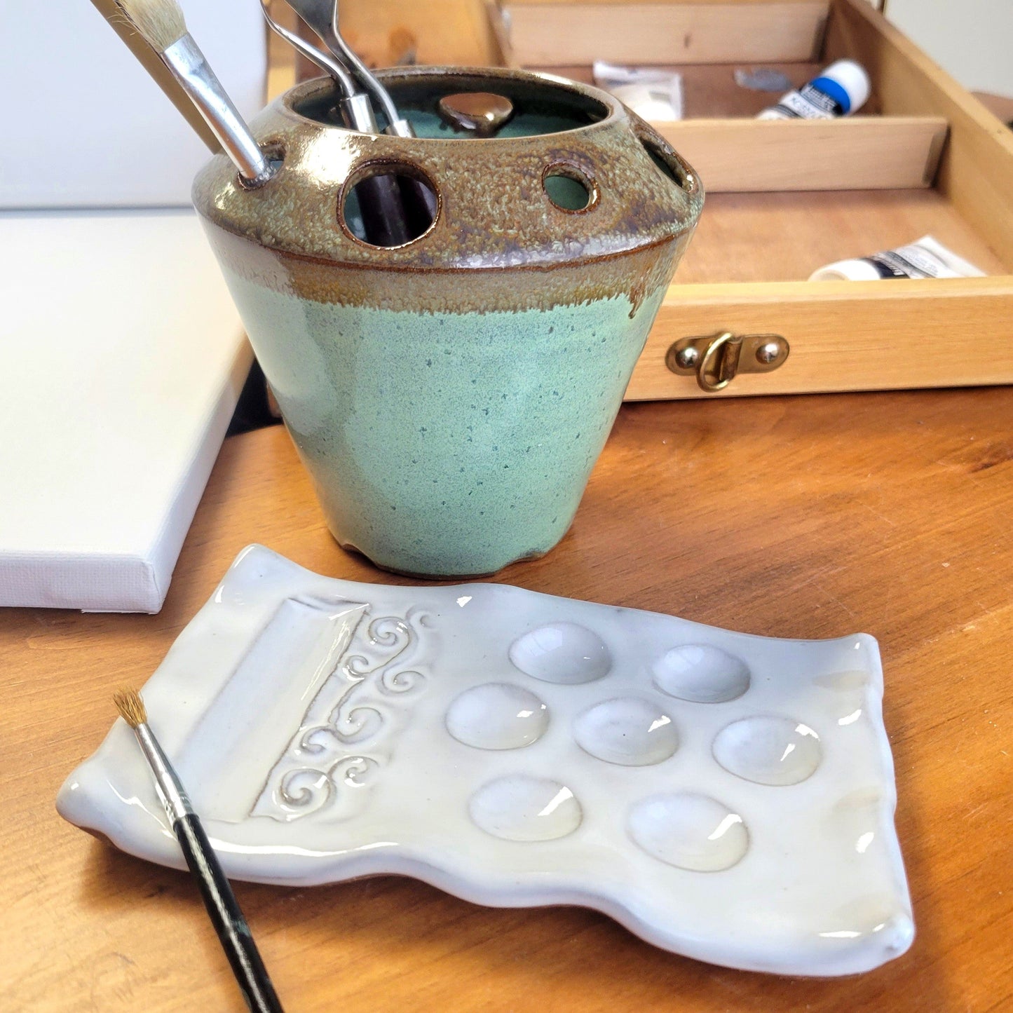 A handcrafted ceramic painting set including a palette and brush organizer caddy a wooden surface with painters canvas, paints and brushes in background.