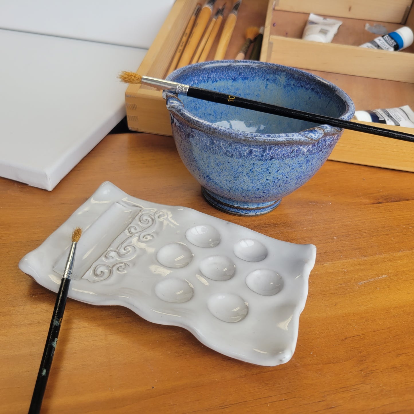 A handcrafted ceramic painting set including a palette and rinse cup, displayed on a wooden surface with brushes and a paintbrush rest. The set is in blue speckle color.