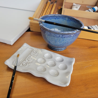 A handcrafted ceramic painting set including a palette and rinse cup, displayed on a wooden surface with brushes and a paintbrush rest. The set is in blue speckle color.