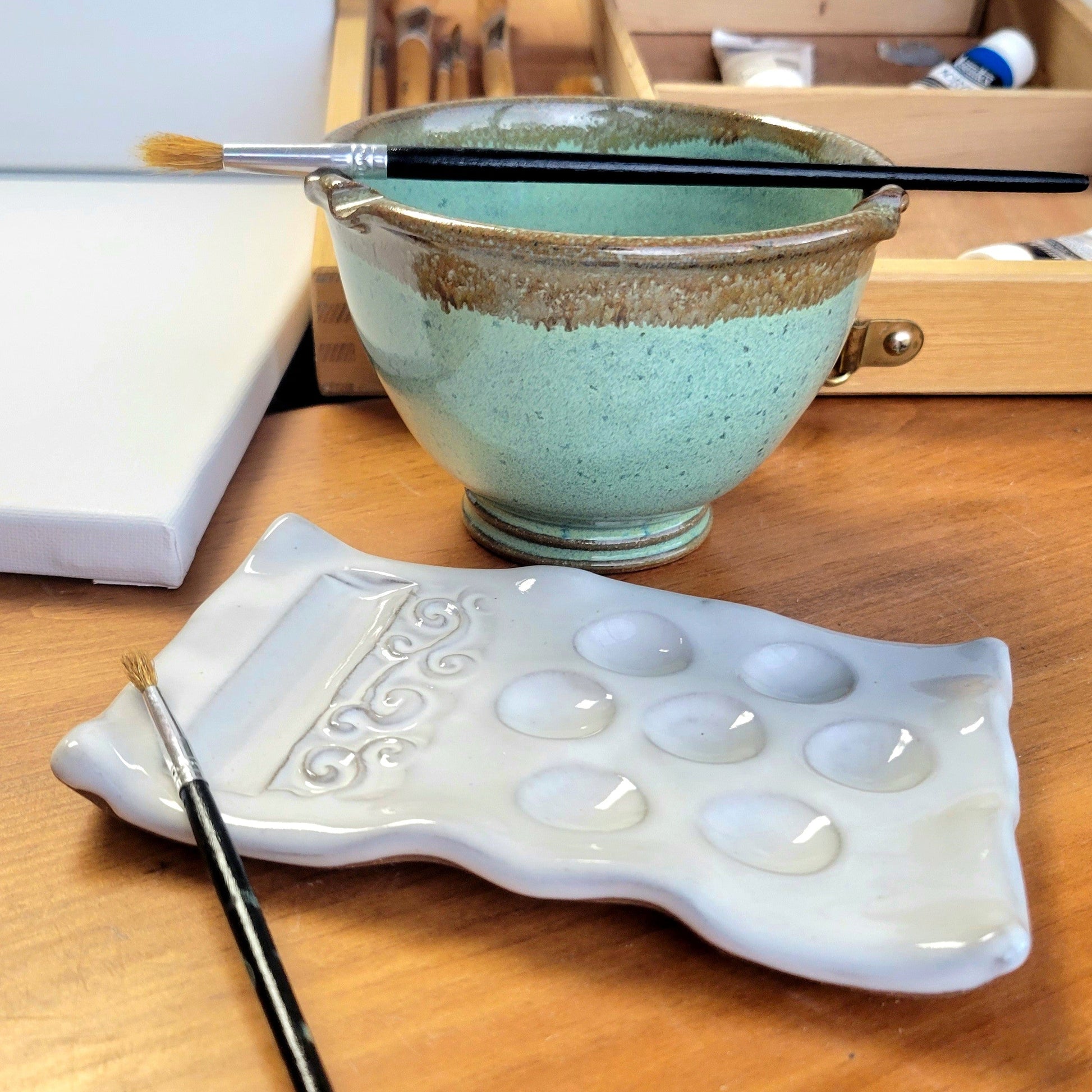 A handcrafted ceramic painting set including a palette and rinse cup a wooden surface with painters canvas, paints and brushes in background.