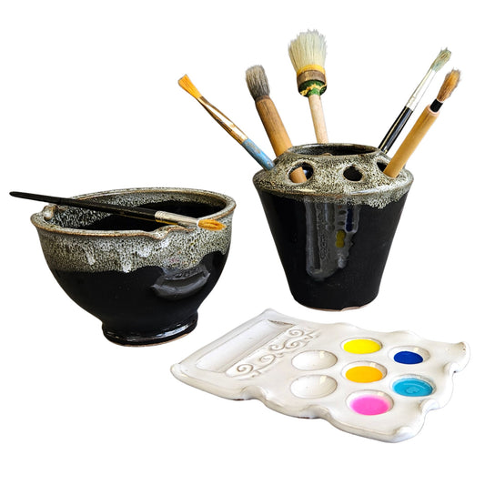 Handmade three-piece ceramic artist set including a black and speckled rinse bowl with brush rest, matching brush holder filled with paintbrushes, and a white ceramic paint palette with colorful paints.