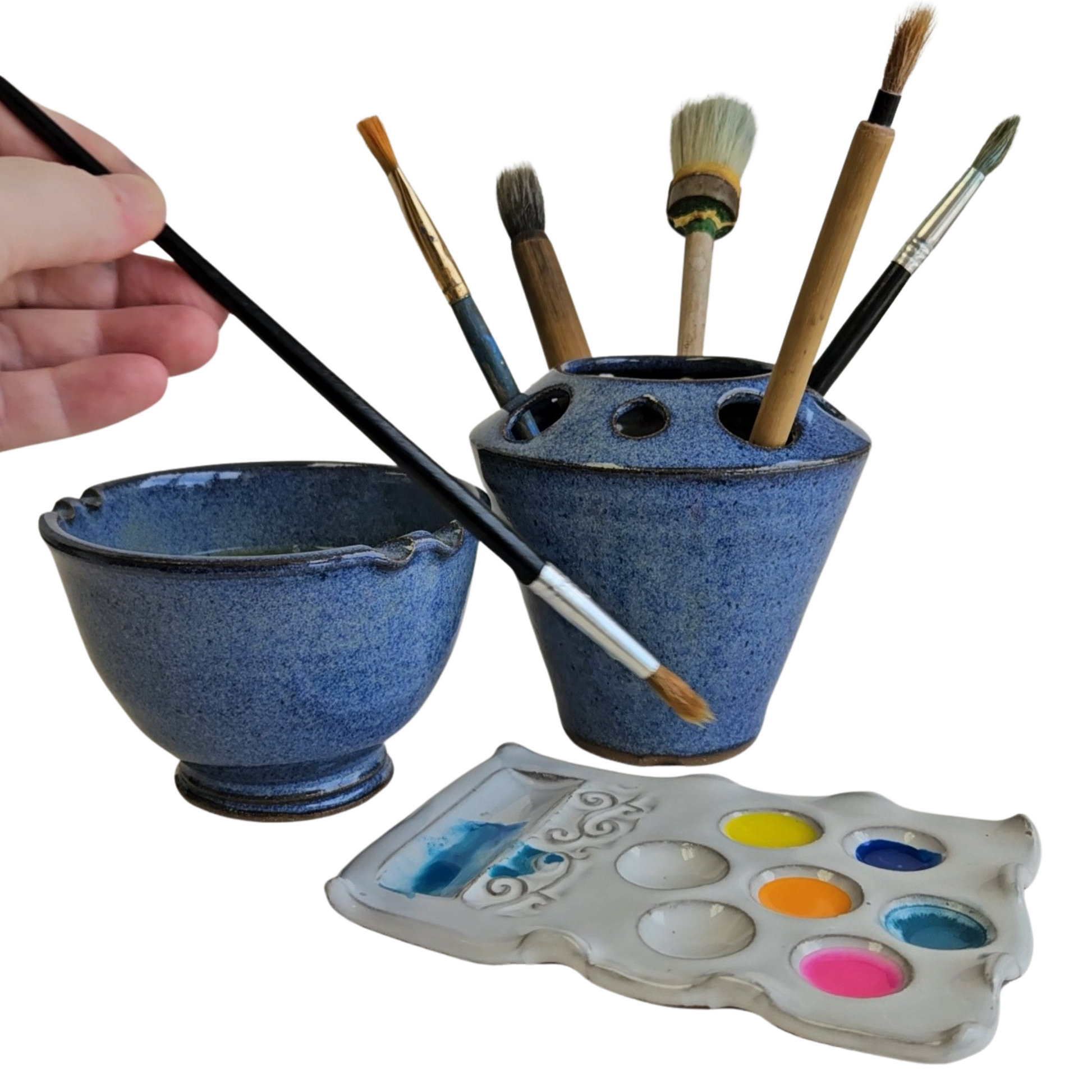 Hand holding a paint brush dipping into a Handmade three-piece blue and white ceramic artist set featuring a rinse bowl with brush rest, a matching brush holder filled with paintbrushes, and a white ceramic paint palette with colorful paints.