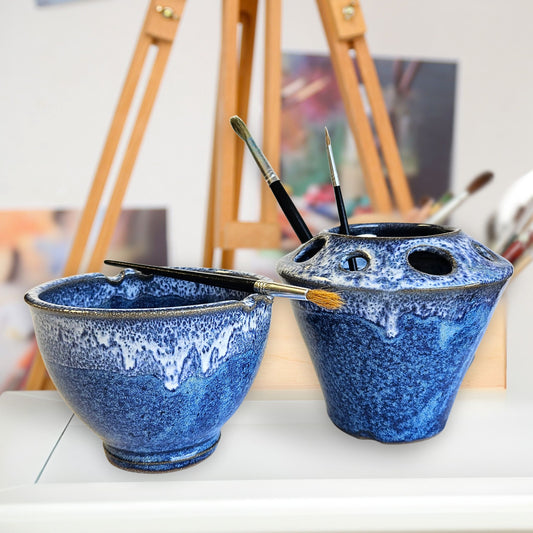Handcrafted blue and white ceramic brush rinse bowl and matching brush caddy with multiple tool slots, holding paintbrushes. The pottery set features a textured drip glaze and is displayed in an artist’s studio with an easel and blurred artwork in the background.