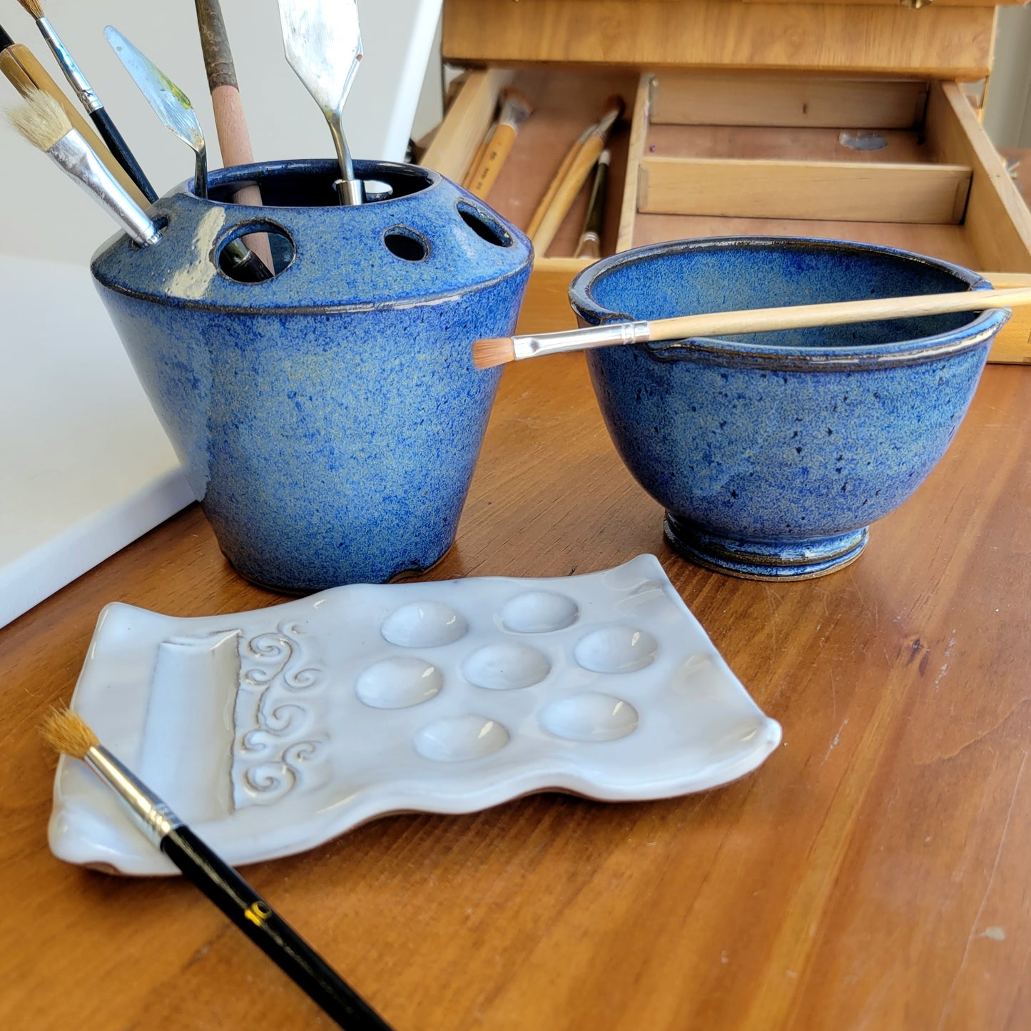 A handcrafted ceramic watercolor painting set including a palette, rinse cup, and caddy, displayed on a wooden surface with art painting supplies in background. The set is primarily blue and white.