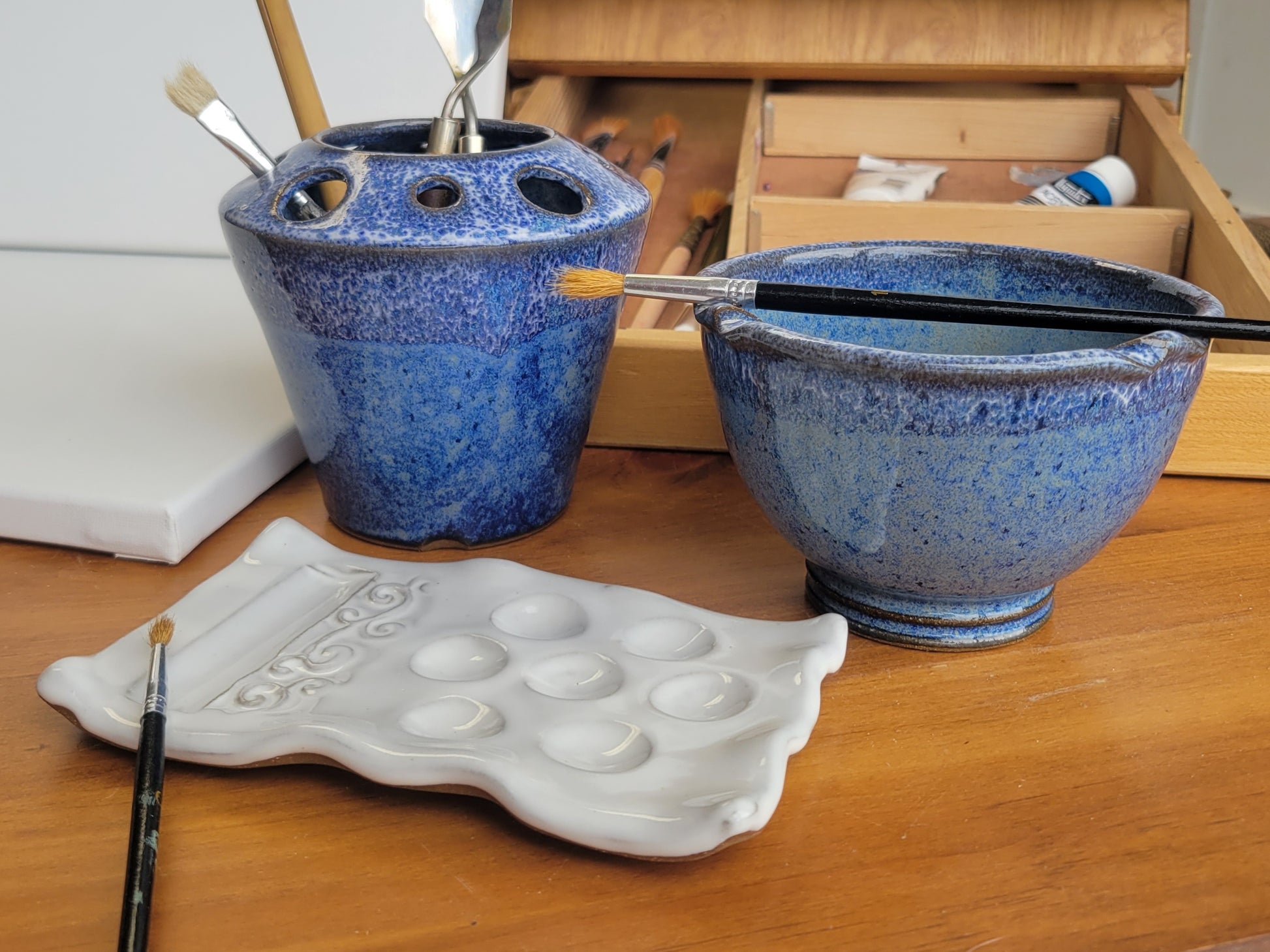 A handcrafted ceramic painting set including a palette, rinse cup, and caddy, displayed on a wooden surface with brushes and a paintbrush rest. The set is in blue speckle color.