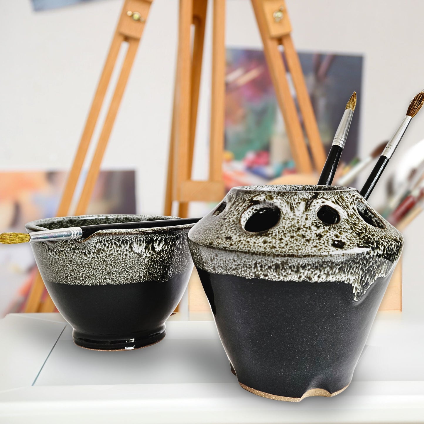 Handmade black and white ceramic paint brush caddy and matching watercolor rinse bowl with multiple tool slots and brush rests, holding paintbrushes. The pottery set features a textured drip glaze and is displayed in an artist’s studio with an easel and blurred artwork in the background.