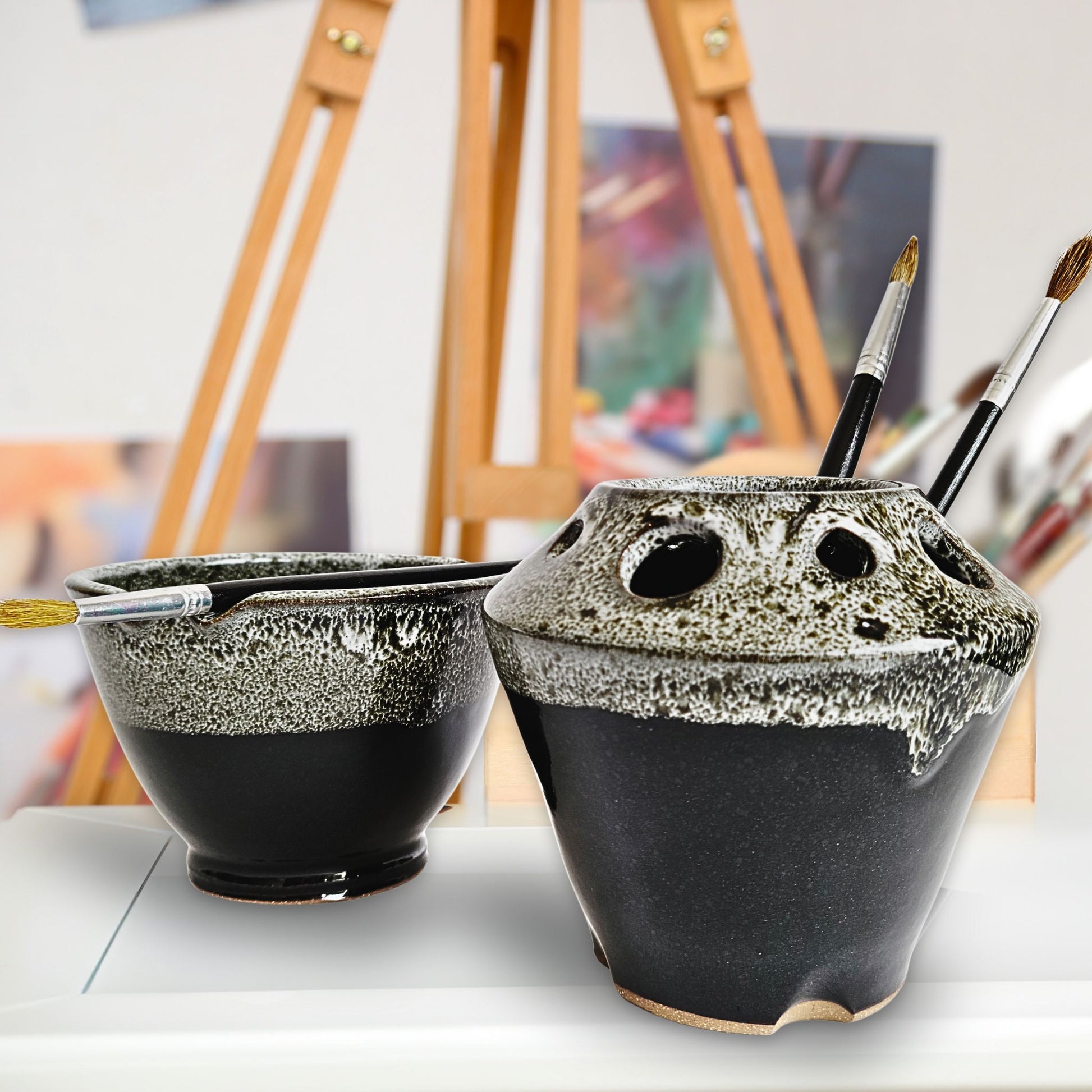 Handmade black and white ceramic paint brush caddy and matching watercolor rinse bowl with multiple tool slots and brush rests, holding paintbrushes. The pottery set features a textured drip glaze and is displayed in an artist’s studio with an easel and blurred artwork in the background.