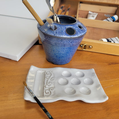 A handcrafted ceramic painting set including a palette and brush caddy, displayed on a wooden surface with brushes and a paintbrush rest. The set is in blue speckle color.