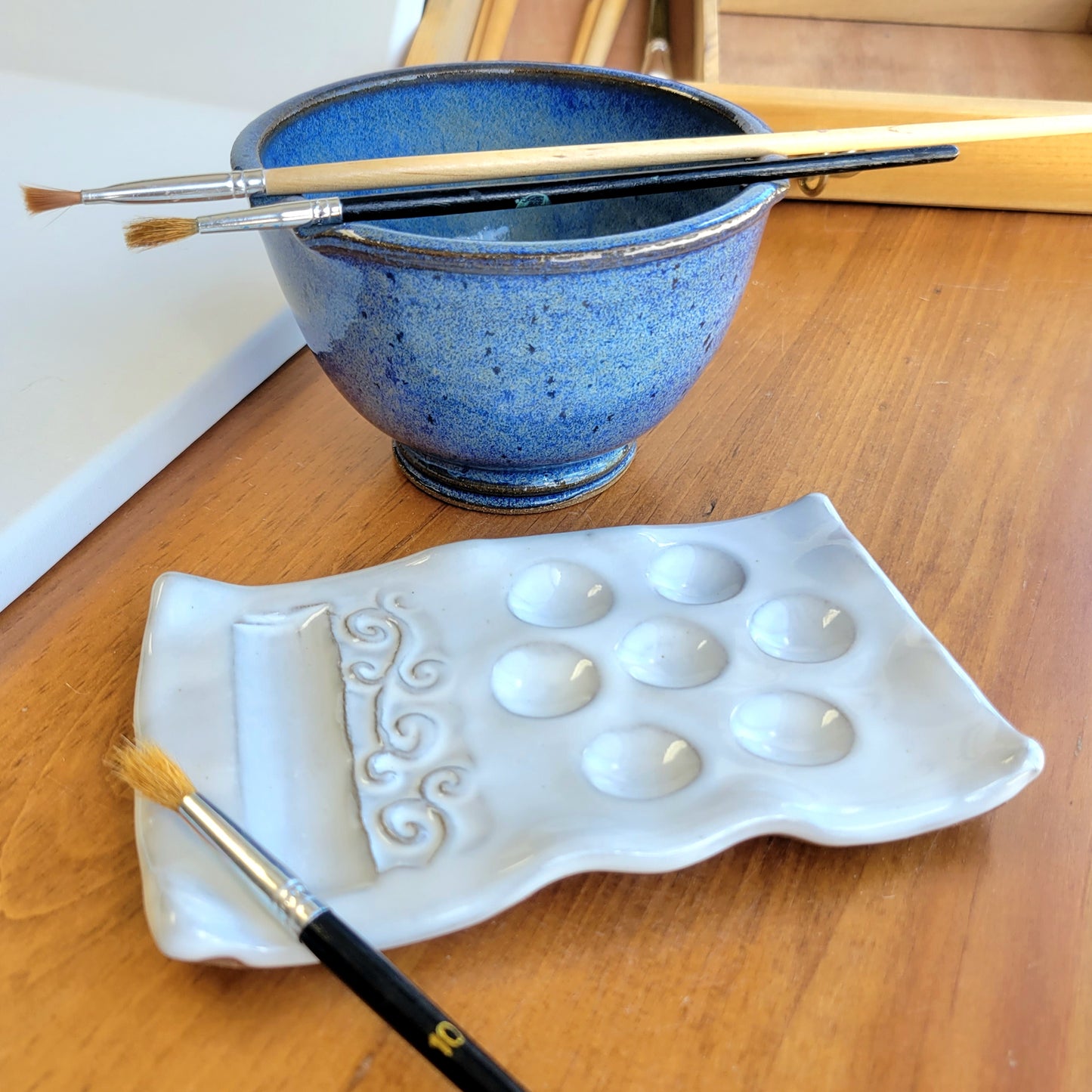 A handcrafted ceramic watercolor painting set including a palette and brush rinse cup displayed on a wooden surface with various paint brushes on it in blue and white.