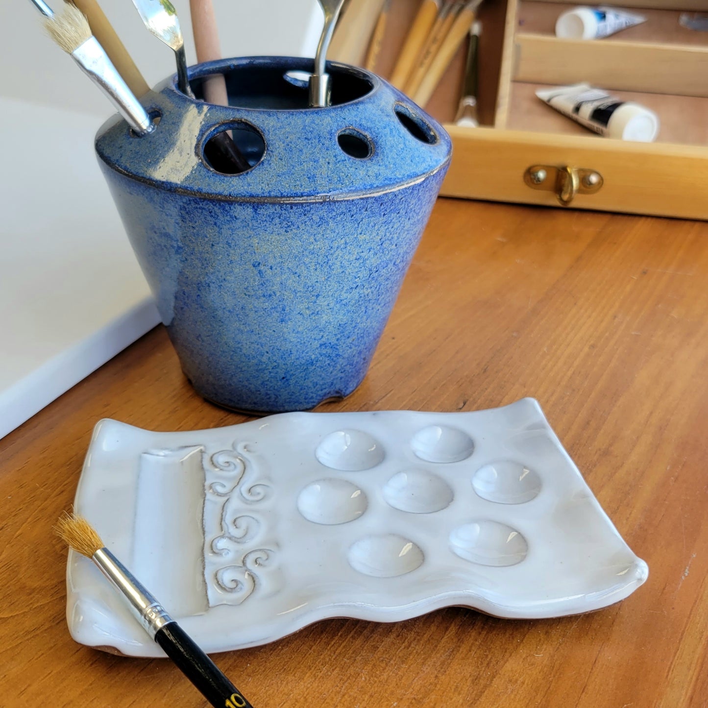 A handcrafted ceramic watercolor painting set including a palette and caddy, displayed on a wooden surface. The set is primarily blue with speckled patterns.