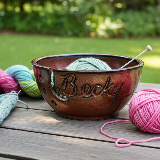 Ceramic bowl personalized with name 'Becky' engraved, containing yarn and a knitting needle on a wooden surface with a blurred green outdoor background.