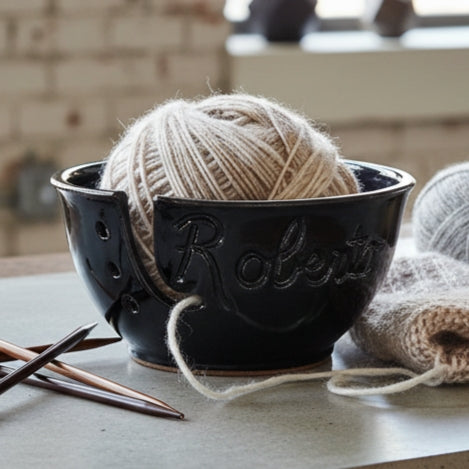 ceramic yarn bowl in black with name 'Roberto' carved into it on a table with knitting needles and project and skeins of brown and grey yarn.