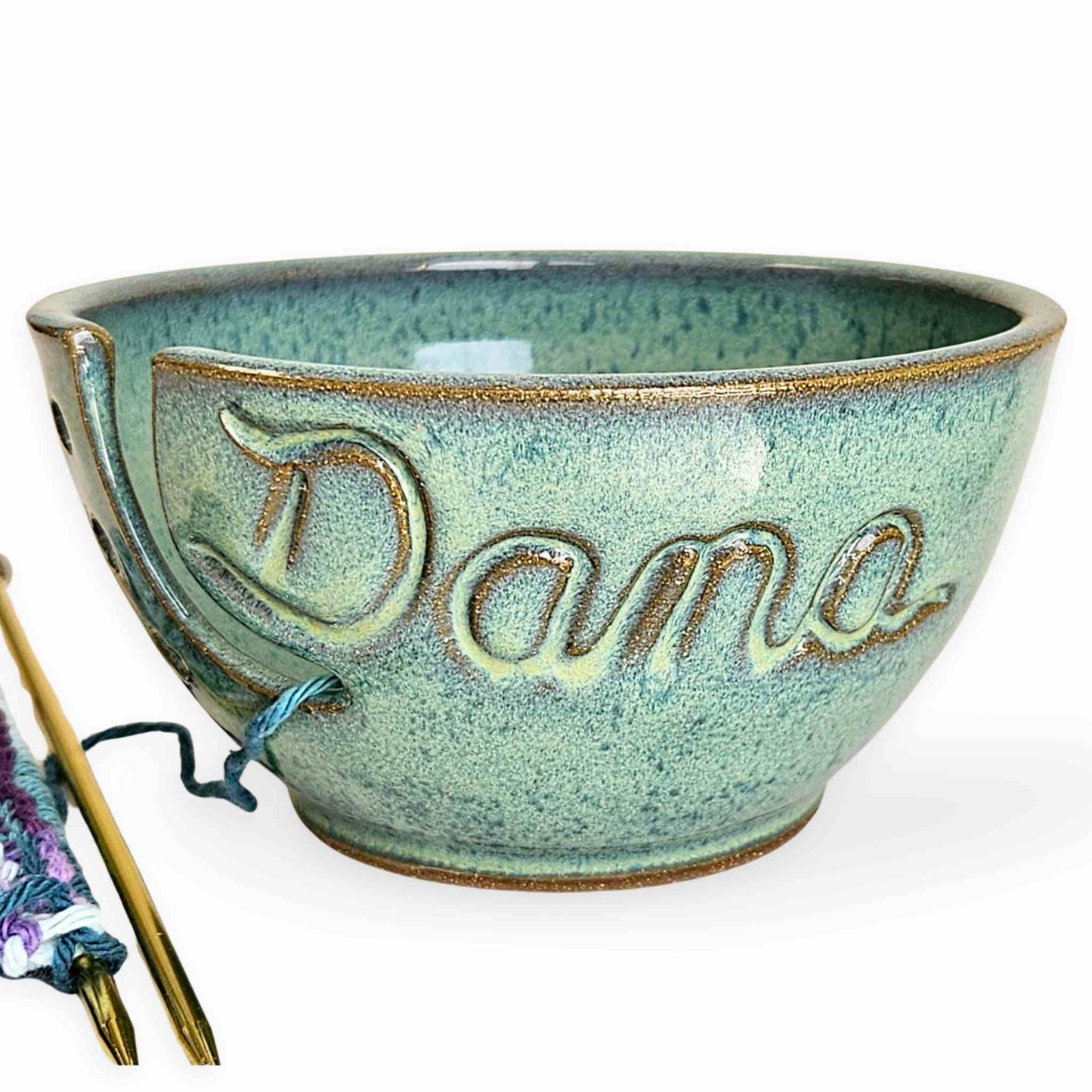 A personalized ceramic yarn bowl with the name 'Dana' carved on the side, containing a skein of blue and purple yarn, alongside a partial view of a knitted piece and knitting needles on a pure white background.
