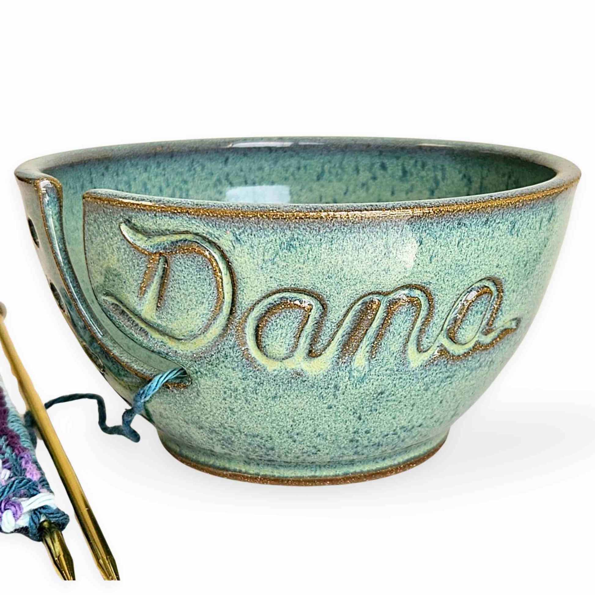 A personalized ceramic yarn bowl with the name 'Dana' carved on the side, containing a skein of blue and purple yarn, alongside a partial view of a knitted piece and knitting needles on a pure white background.