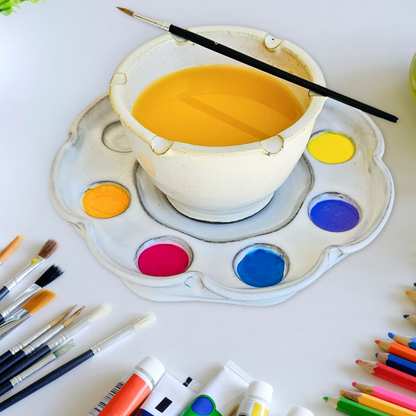 Paint set in a floral shape with paints in the wells and a water rinse bowl with brush rests and a paint brush resting on top on an artists studio table with paints pencils and brushes