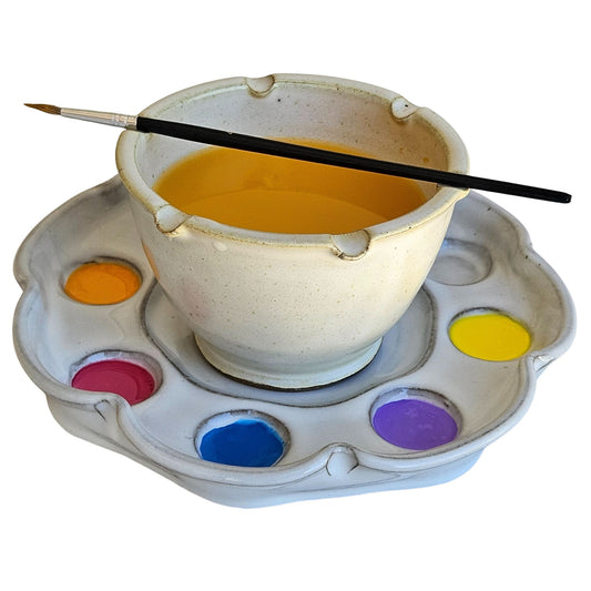 Handcrafted ceramic paint rinse cup with brush rests on rim and paint inside on a matching palette with paint color swatches.