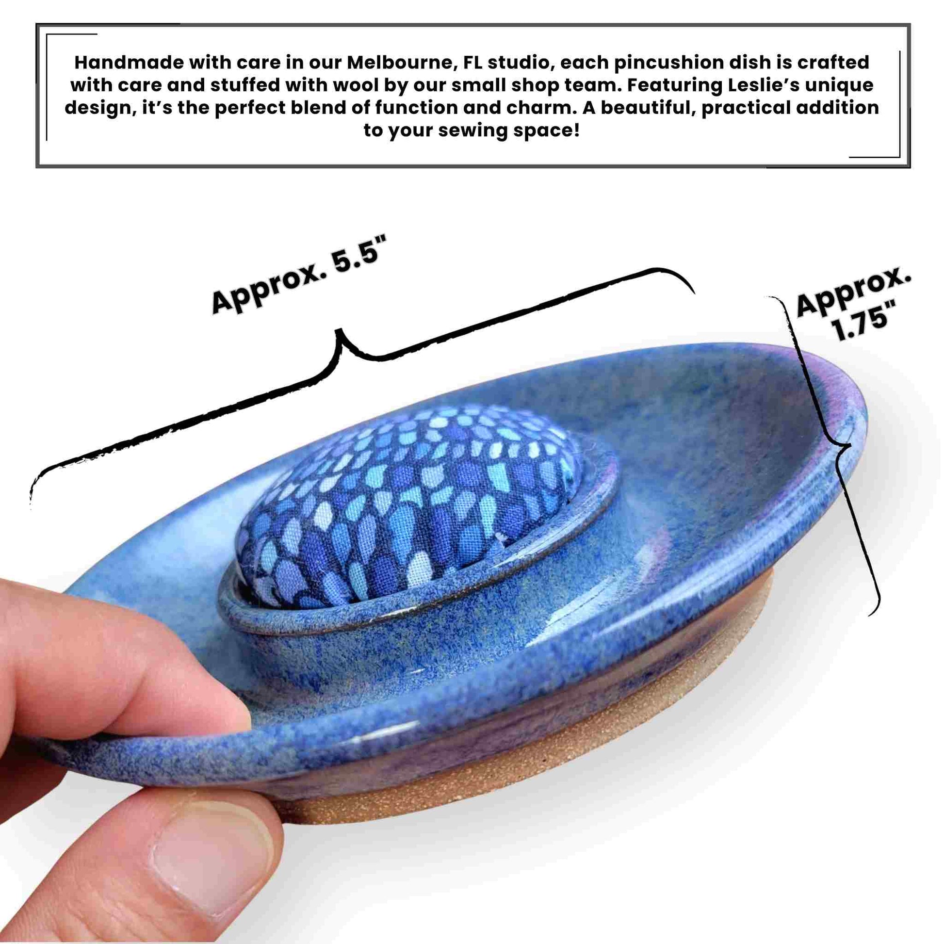 Handmade ceramic sewing caddy with a blue patterned fabric pincushion, displayed in a glossy blue dish. Includes silver scissors, yellow measuring tape, pink thread spool, and a metal thimble, styled on a wooden sewing table with a softly blurred vintage sewing machine in the background.