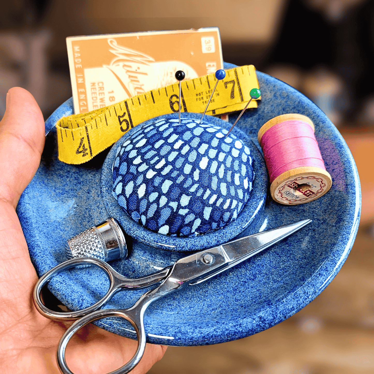 Handmade ceramic sewing caddy with a blue patterned fabric pincushion, displayed in a glossy blue dish. Includes silver scissors, yellow measuring tape, pink thread spool, and a metal thimble, styled on a wooden sewing table with a softly blurred vintage sewing machine in the background.