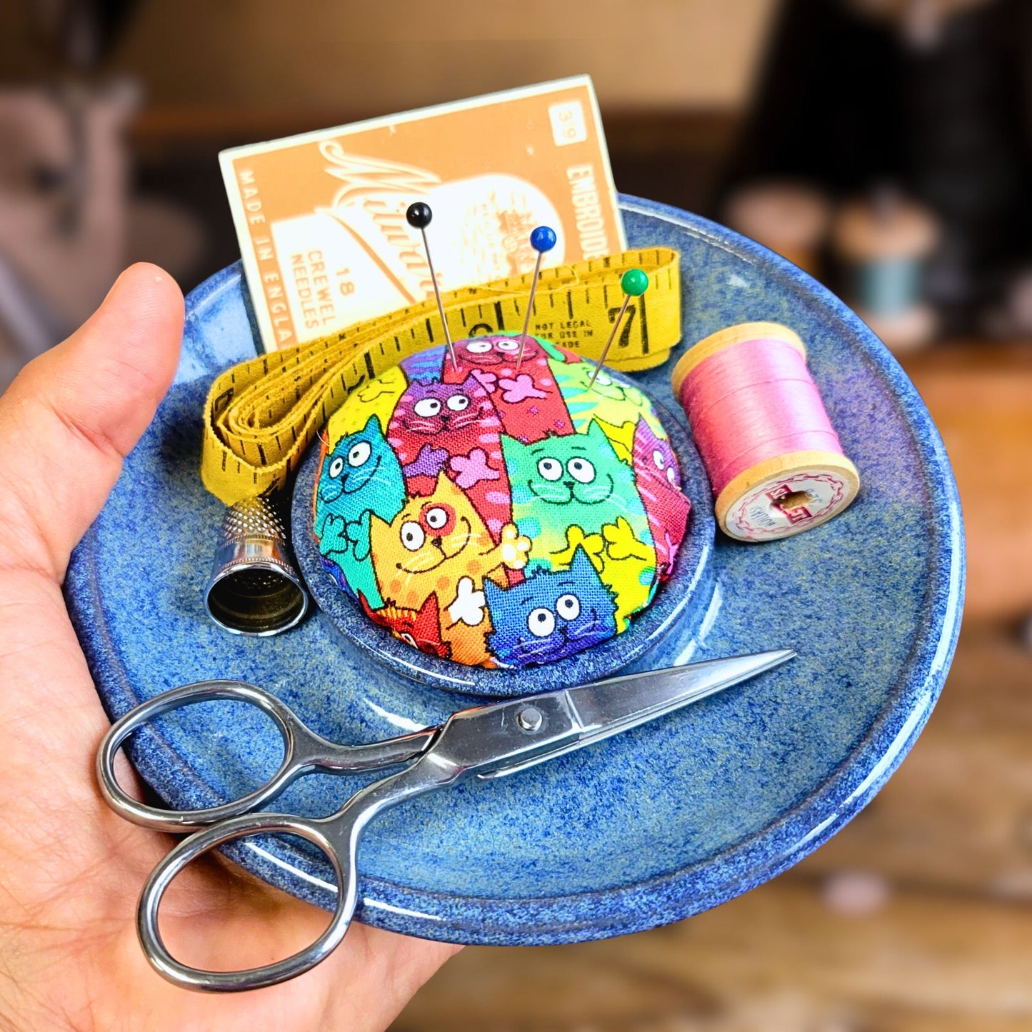 Handmade ceramic sewing caddy with a rainbow cat fabric pincushion, displayed in a glossy blue dish. Includes gold stork scissors, yellow measuring tape, pink thread spool, and silver thimble, styled on a warm wooden sewing table with a softly blurred vintage sewing machine in the background.