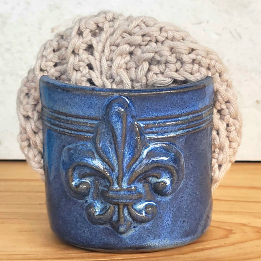 Front view of ceramic blue sponge keeper with a decorative fleur-de-lis design holding a crochet cotton yarn scrubber sponge. Perfect for kitchen counter organization. 