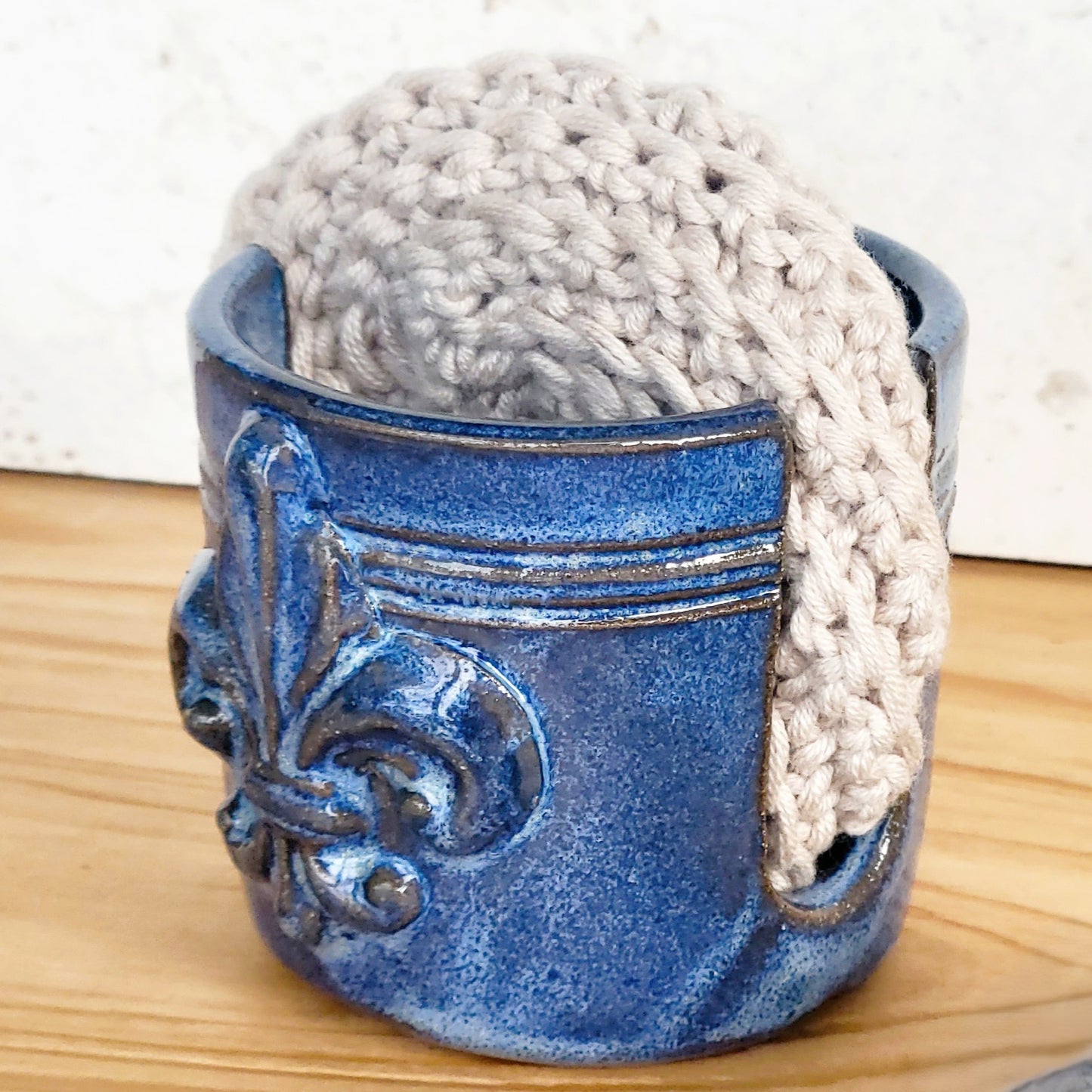 Side view of ceramic blue sponge keeper with a decorative fleur-de-lis design holding a crochet cotton yarn scrubber sponge. Perfect for kitchen counter organization. 