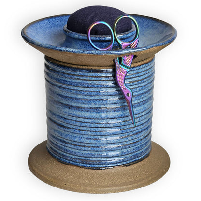 Blue ceramic sewing caddy shaped like an oversized vintage spool with navy pincushion, bobbin holder and scissor compartment with inner storage for sewing and mending supplies.