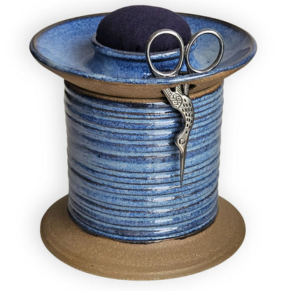 Blue ceramic sewing caddy shaped like an oversized vintage spool with navy pincushion, bobbin holder and scissor compartment with inner storage for sewing and mending supplies.
