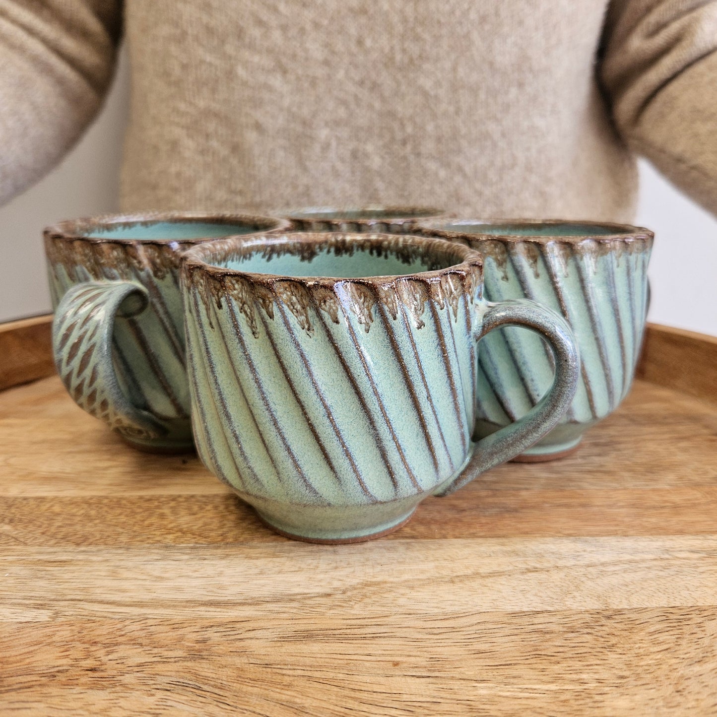 Set of four ceramic mugs with diagonal striped design in a soft green and brown drip glaze on a wooden surface held by a person in a cozy sweater
