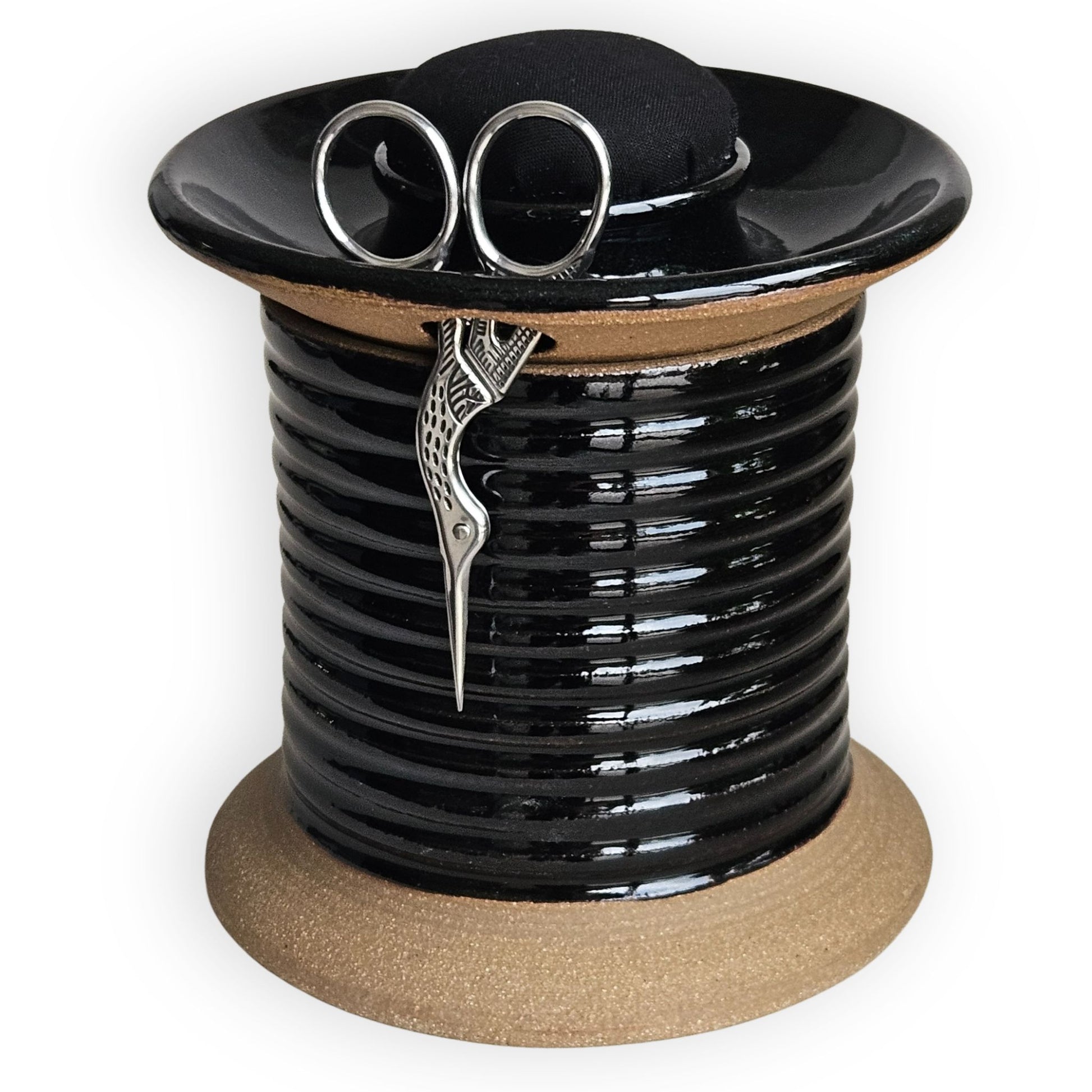 Tailors sewing project notions holder shaped like oversized spool for runway fashion alterations in black ceramic with modern black pincushion topper, crane scissors, and storage compartments for bobbins and sewing notions.