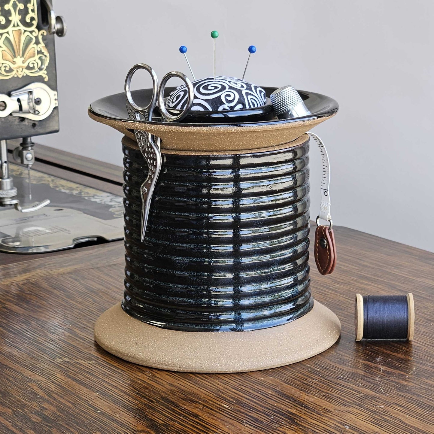 A black oversized spool-shaped  ceramic sewing caddy with a pincushion topper, removable lid, and slots for storing sewing tools such as scissors, bobbins, thimble, and tape measure, displayed on an antique Singer sewing machine next to a spool of thread.