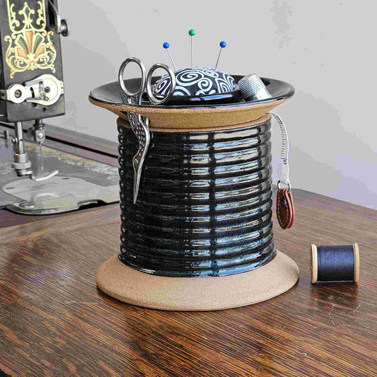 A black oversized spool-shaped  ceramic sewing caddy with a pincushion topper, removable lid, and slots for storing sewing tools such as scissors, bobbins, thimble, and tape measure, displayed on an antique Singer sewing machine next to a spool of thread.