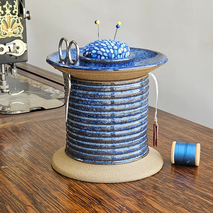 A handmade ceramic sewing station caddy in the shape of an oversized spool, featuring a blue and brown pattern, with a pincushion lid and sewing tools arranged on top.