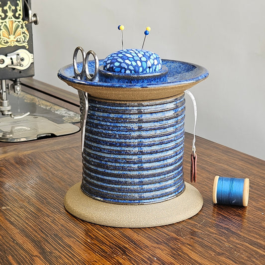 A handmade ceramic sewing station caddy in the shape of an oversized spool, featuring a blue and brown pattern, with a pincushion lid and sewing tools arranged on top.