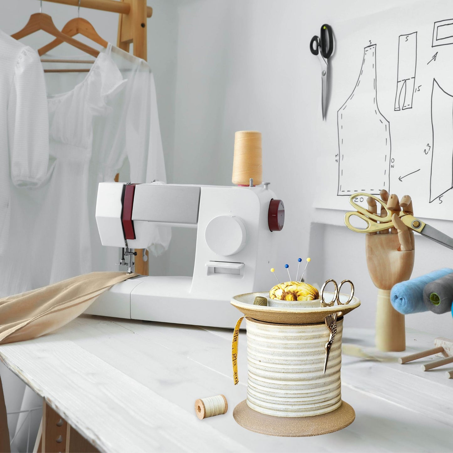 A sewing room table with spool shaped caddy filled with sewing notions, pins and scissors on a table with sewing machine and garments and patterns in the background.
