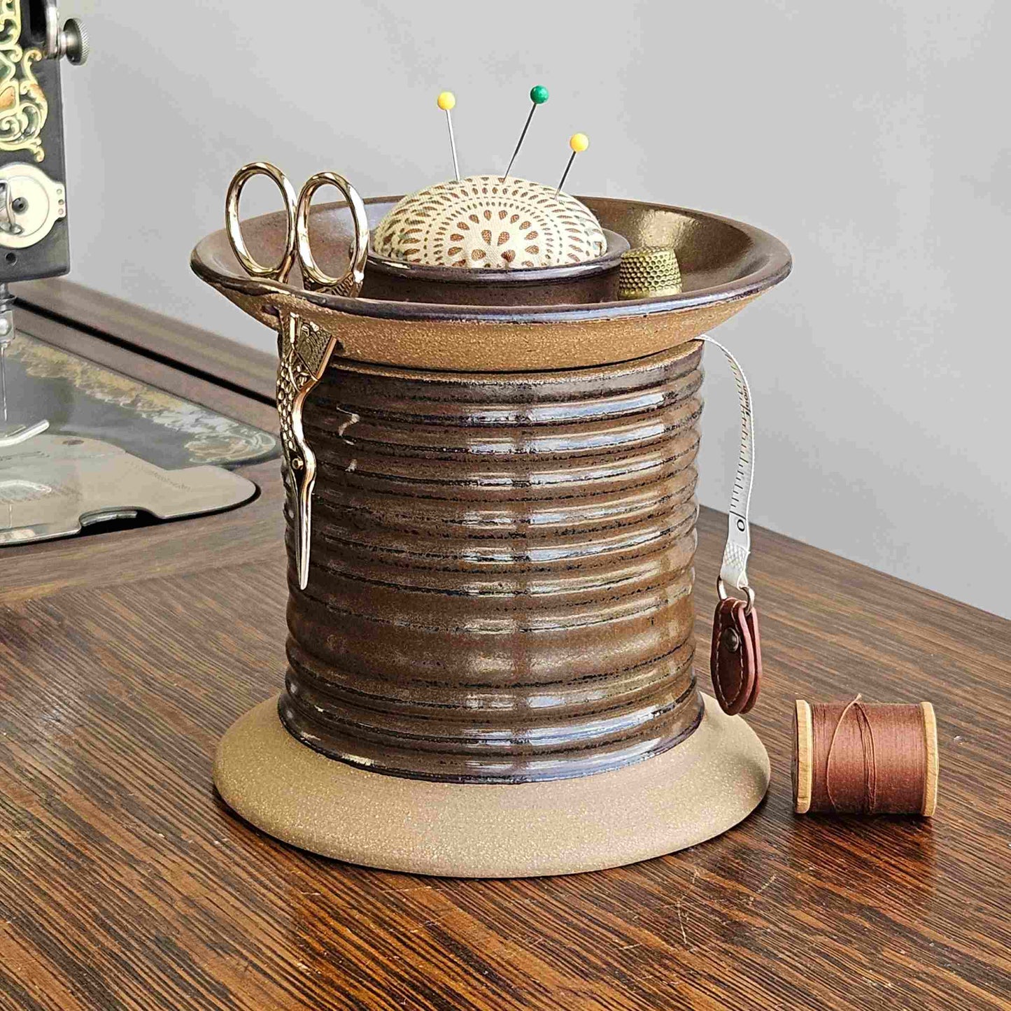A ceramic sewing organizer caddy shaped like an oversized spool with removable lid, scissor holder and pincushion topper. It is placed on a wooden surface next to an old sewing machine with a few sewing accessories like scissors and bobbins stored on it.